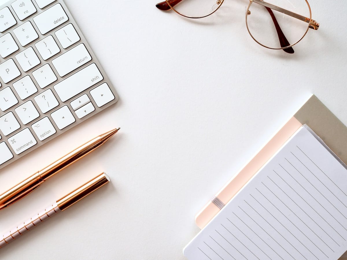 A keyboard, pen, notebook and glasses on a white desk