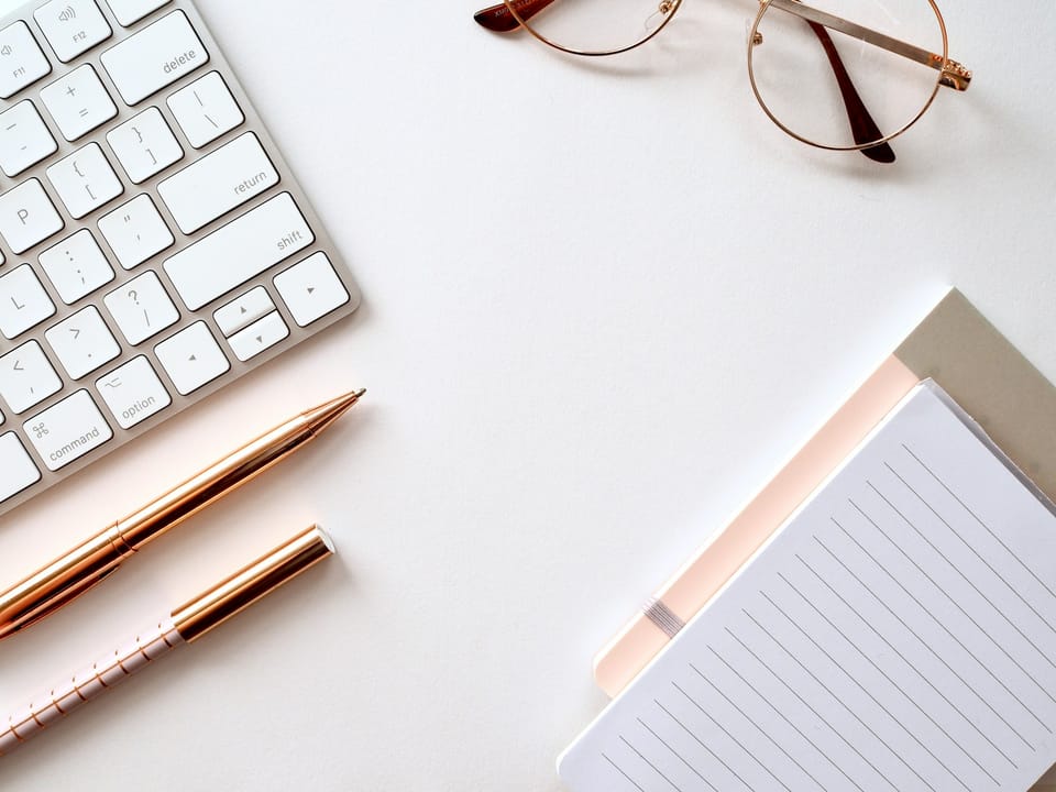 A keyboard, pen, notebook and glasses on a white desk