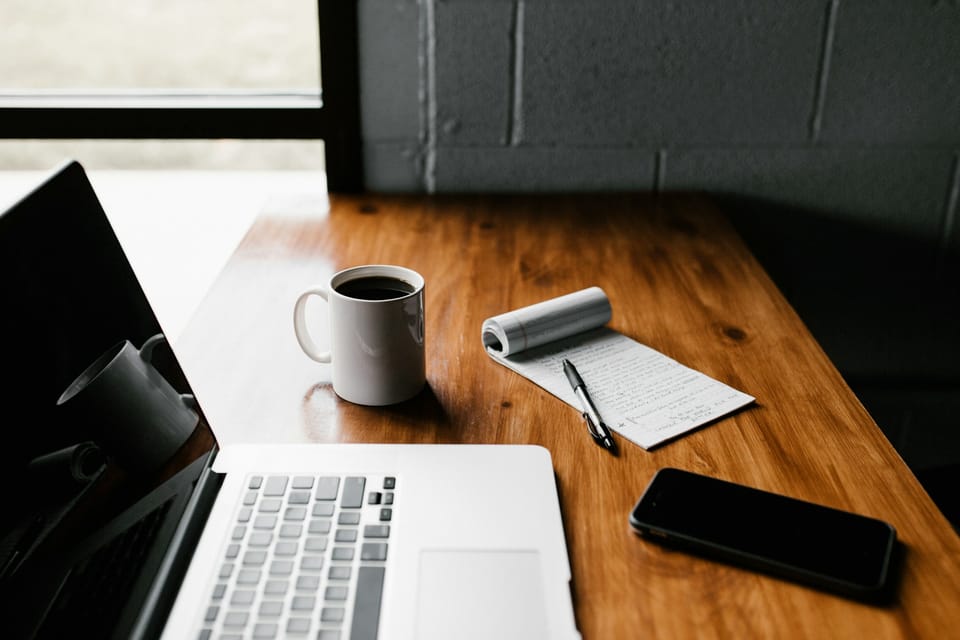 A laptop, mug, notepad and phone on a wooden desk